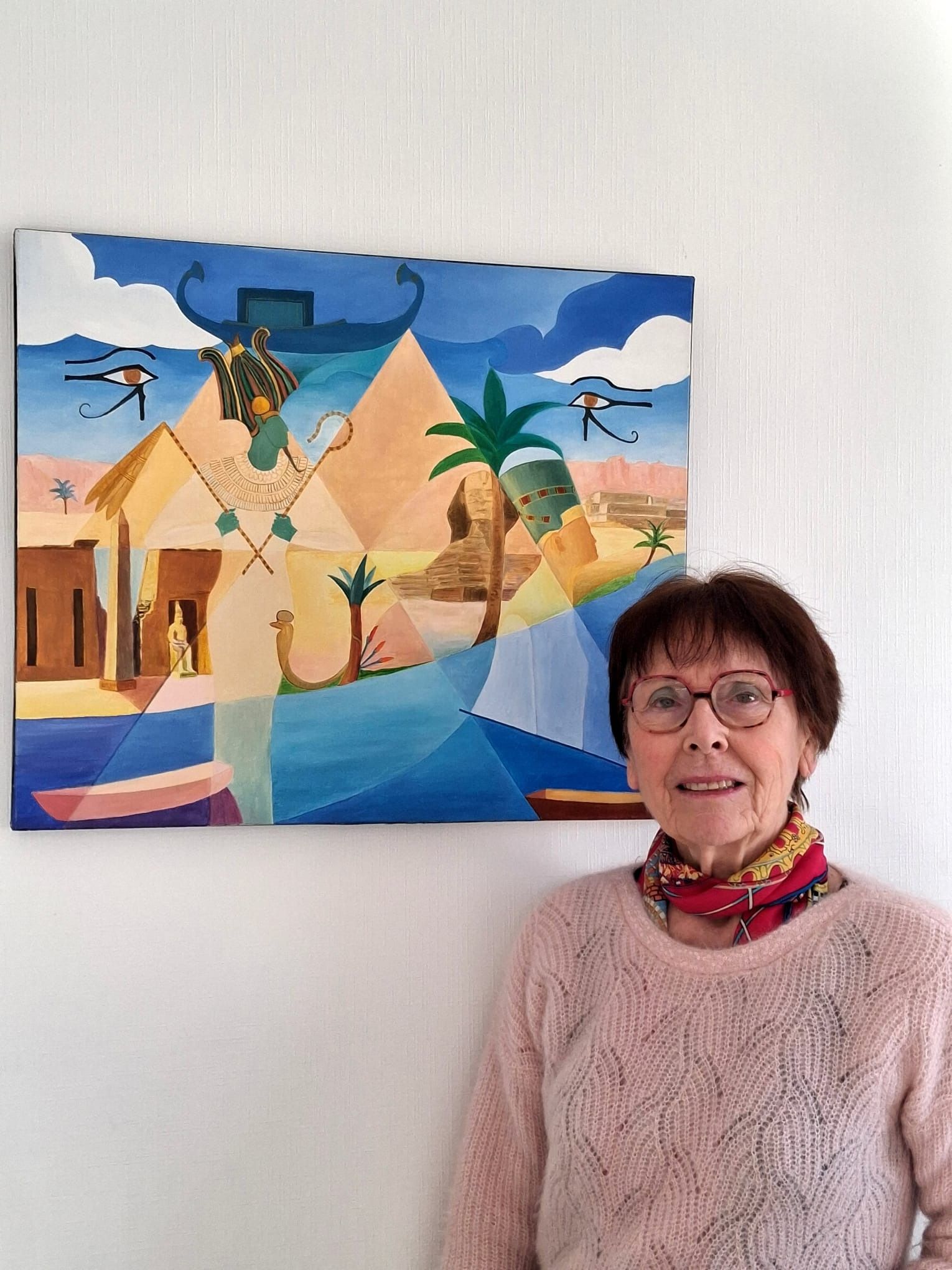 Monique Amiot, souriante, en pull rose et foulard coloré, devant l'une de ses toiles : une Égypte imaginaire aux pyramides et aux palmiers.